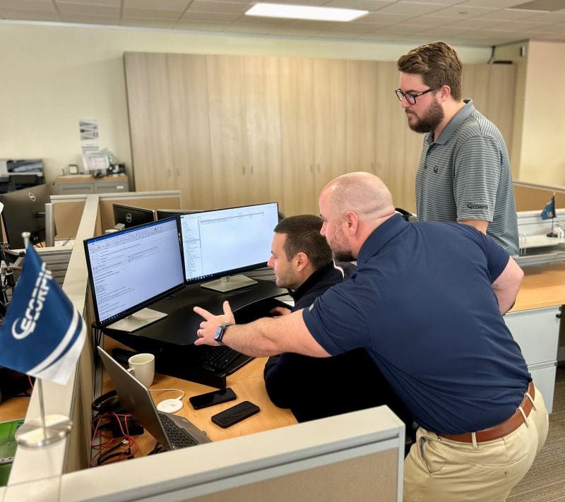 Three men at a cubicle looking at the computer, one is pointing to the screen.