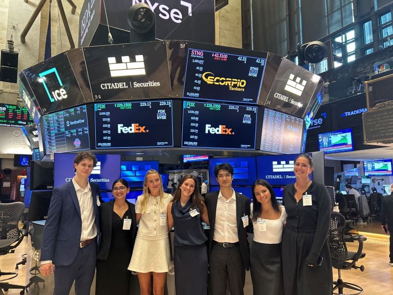 Group of summer interns standing on the floor of the NYSE, below the screens displaying the Scorpio Tankers stock.